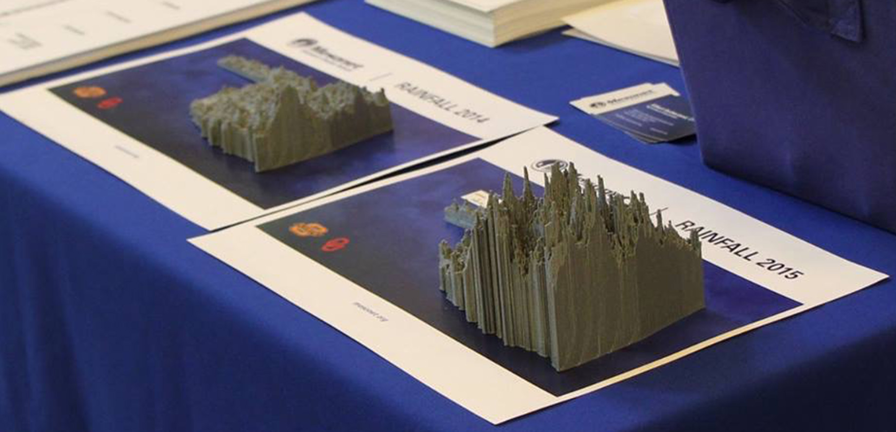Two gray, 3-d printed models of rainfall sitting on a blue tablecloth at a public exhibit hall.
